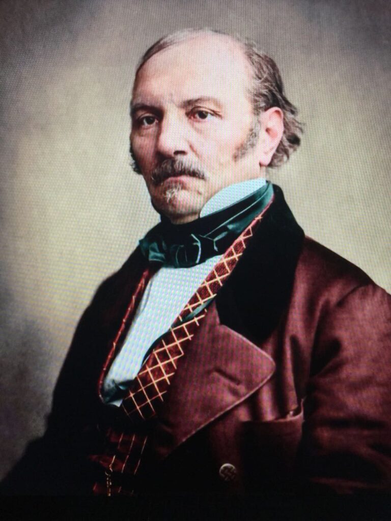 Retrato de un hombre con expresión seria, vistiendo saco burdeos, chaleco con patrón geométrico y corbata verde, sobre un fondo neutro.