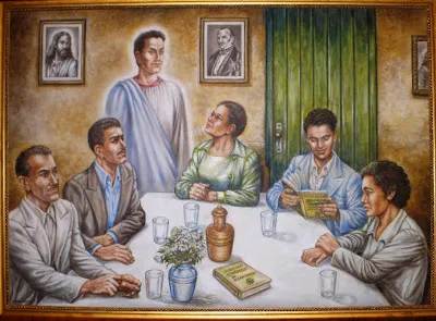 Representación artística de una reunión espiritista, con cinco personas sentadas alrededor de una mesa leyendo y conversando, mientras un espíritu luminoso aparece entre ellos. En la pared hay retratos enmarcados y libros espiritistas sobre la mesa.