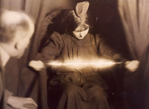 Médium de efecto físico manifestando energía espiritual, con rayos de luz visibles entre sus manos, mientras una persona observa atentamente.